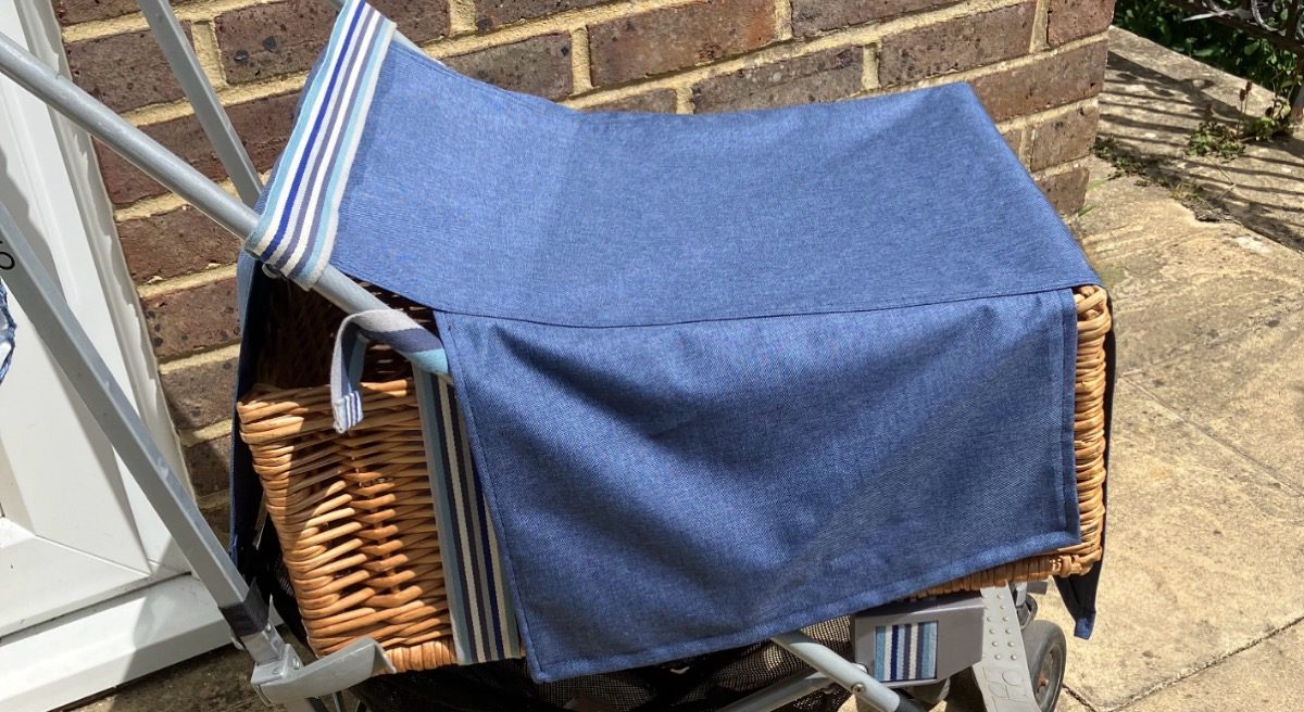 An umbrella-folding pushchair has been adapted into a shopping trolley with the addition of a wicker basket. It has a blue waterproof cover.