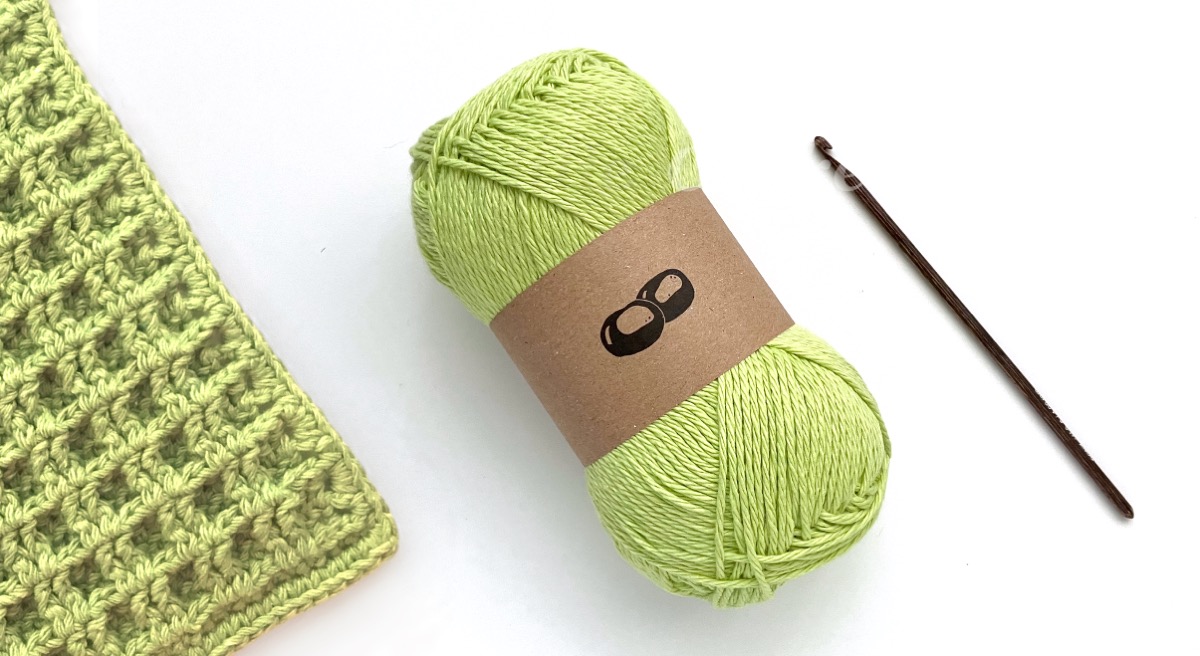 A ball of vibrant lime green recycled cotton yarn, a dishcloth made from the yarn and a wooden crochet hook
